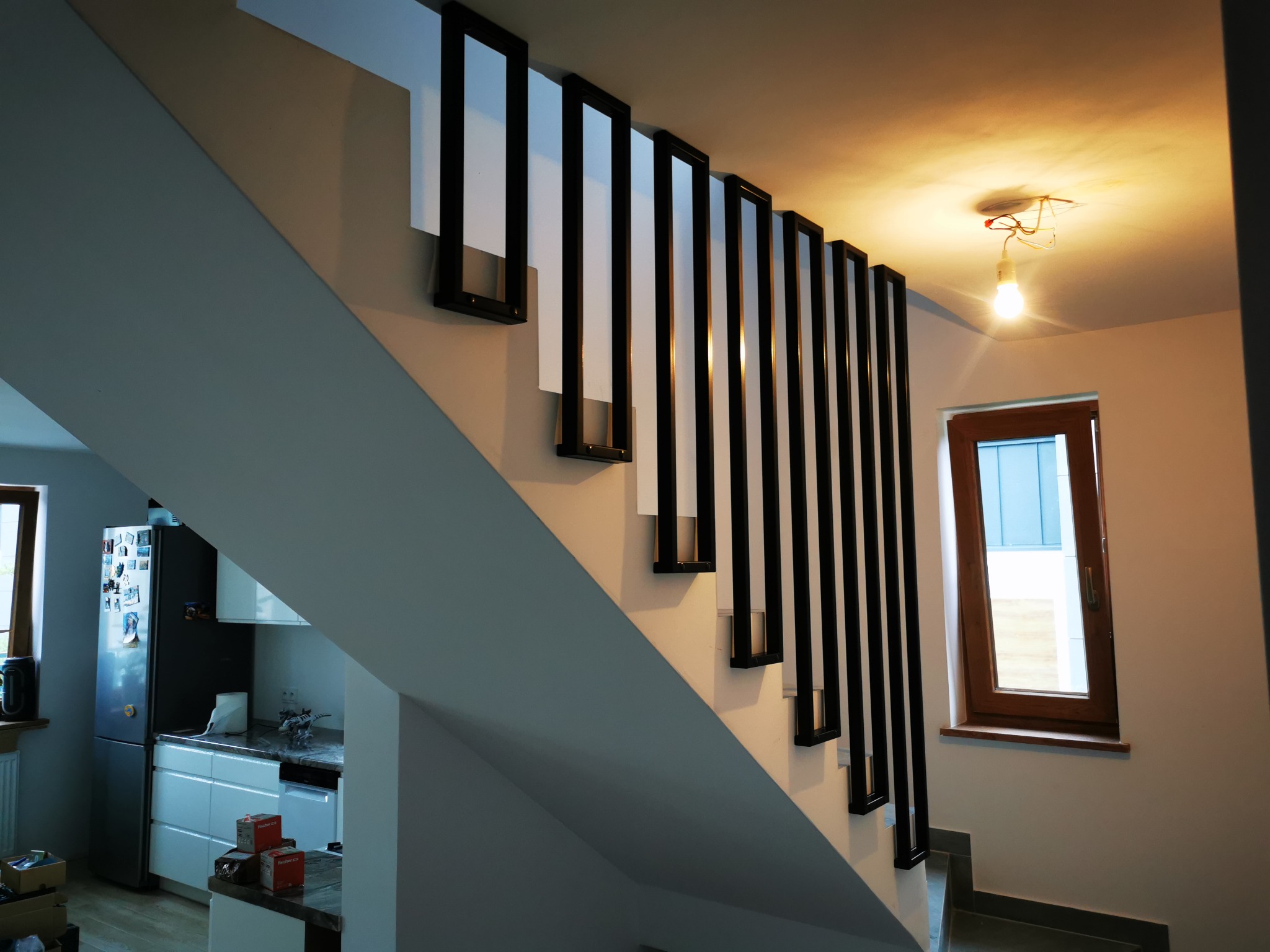 Nowoczesne schody z białą konstrukcją i czarnymi, pionowymi elementami dekoracyjnymi, widoczne z dołu, z niedokończonym oświetleniem sufitowym i oknem w tle.