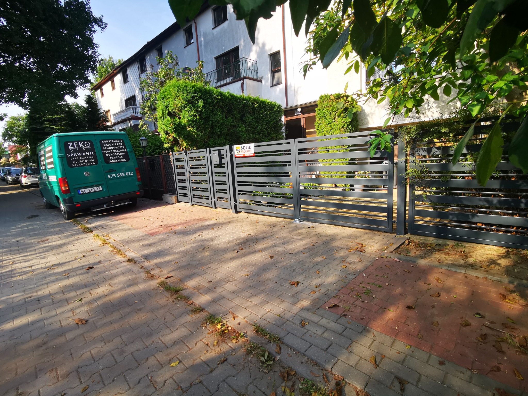 Nowoczesna, pozioma brama wjazdowa z szarych, metalowych profili, z tabliczką 'Solid Security', przed budynkiem mieszkalnym i zaparkowanym zielonym vanem firmy z branży metalowej.