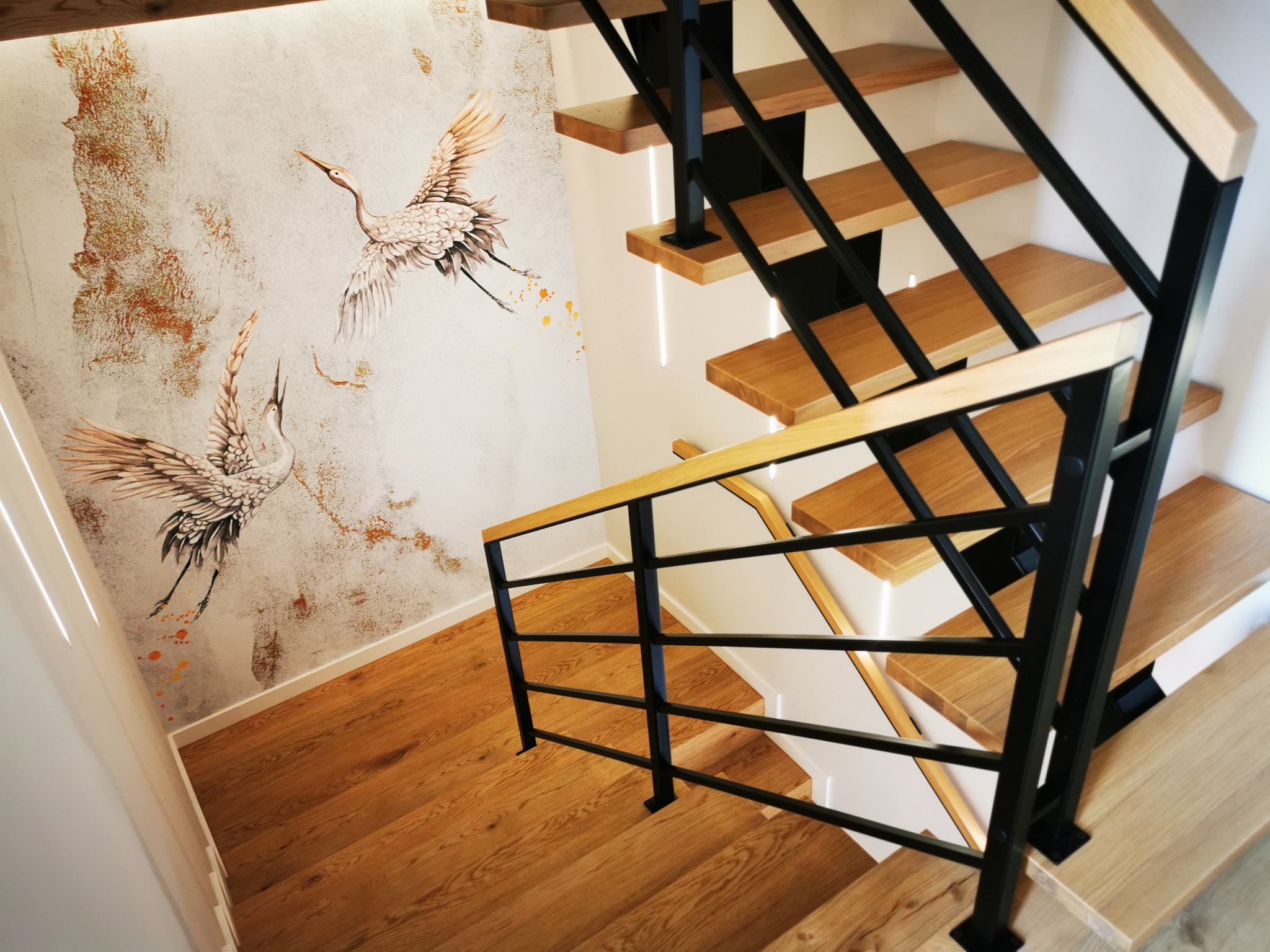 Drewniane schody z czarną metalową balustradą i drewnianymi poręczami, na ścianie dekoracyjny motyw żurawi.