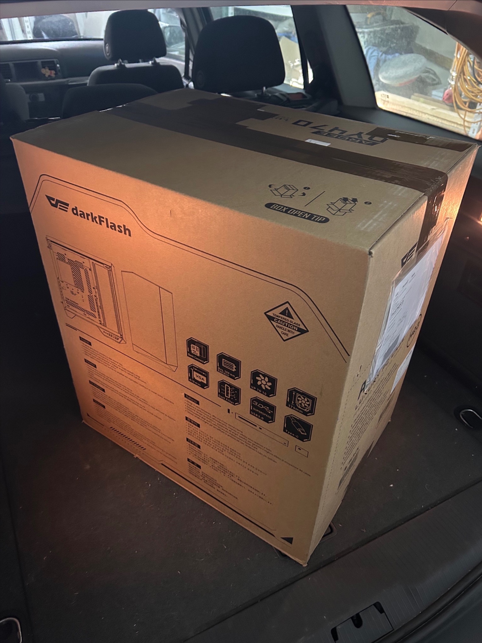 Pudło obudowy komputera Darkflash w bagażniku samochodu. Widoczne specyfikacje techniczne i ostrzeżenia na opakowaniu. Gotowy do transportu, złożony komputer.