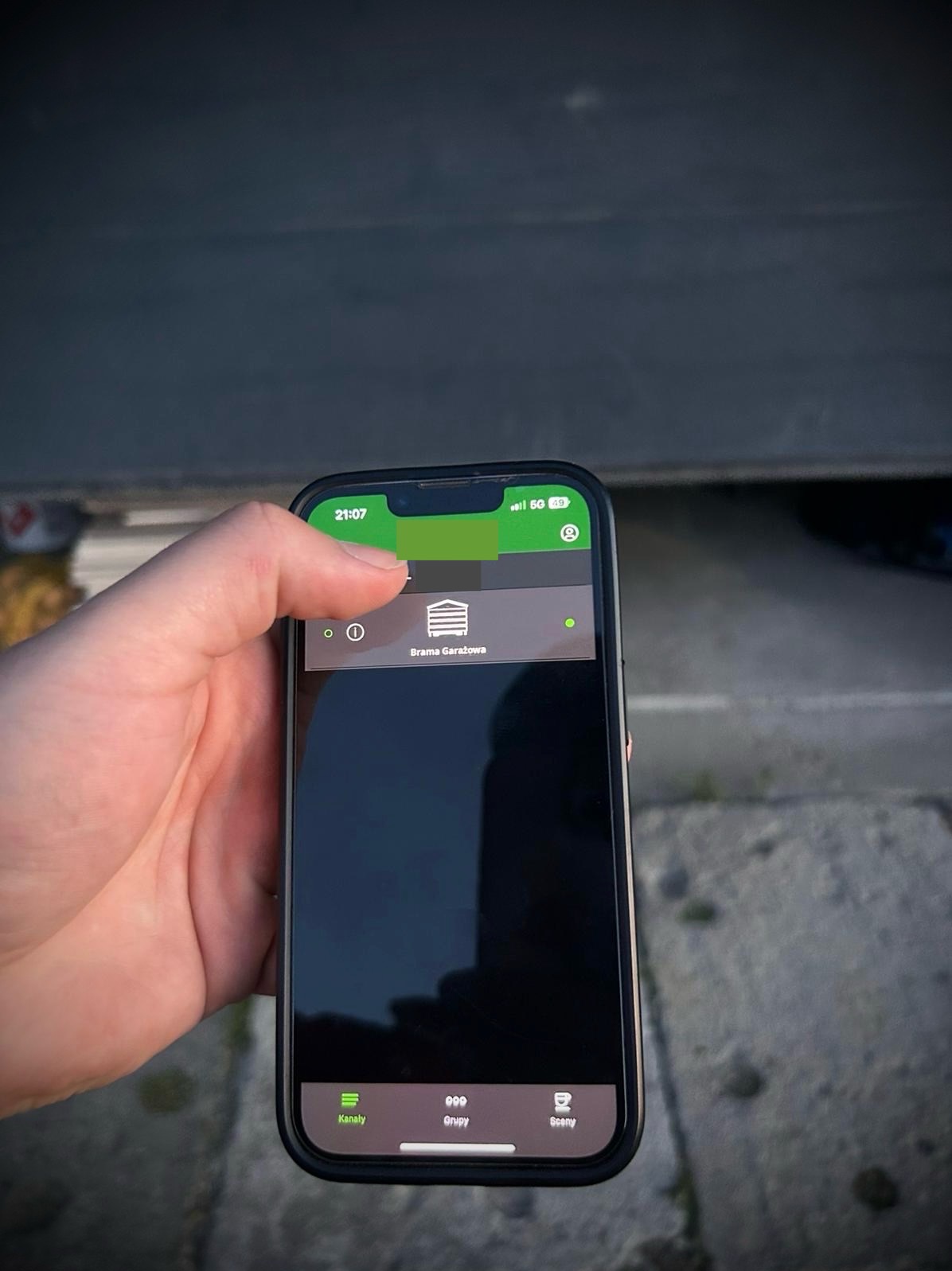 Dłoń trzyma smartfon z aplikacją do sterowania bramą garażową. Widoczny interfejs aplikacji z ikoną bramy i napisem 'Brama Garażowa'.