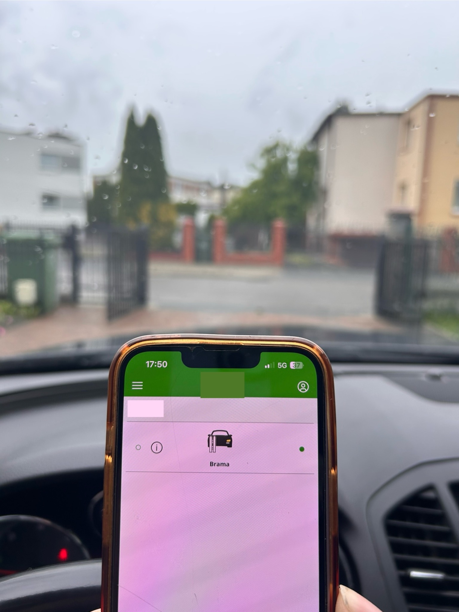 Aplikacja do sterowania bramą automatyczną na smartfonie, widok z wnętrza samochodu na bramę wjazdową w tle, pochmurny dzień.