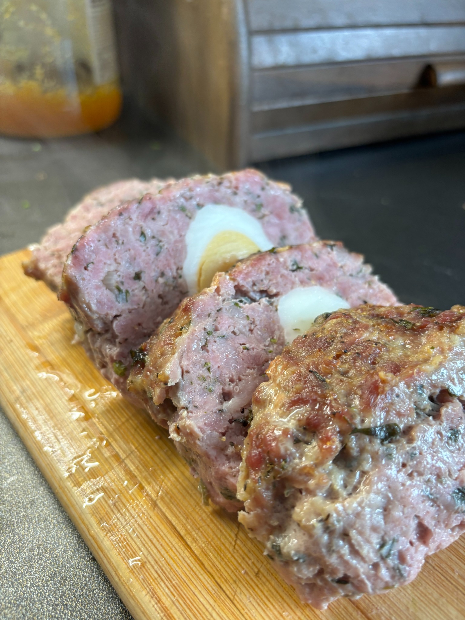 Krojony mięsny pasztet z jajkiem na twardo w środku, ułożony na drewnianej desce do krojenia, z widocznymi ziołami i przyprawami w mięsie.