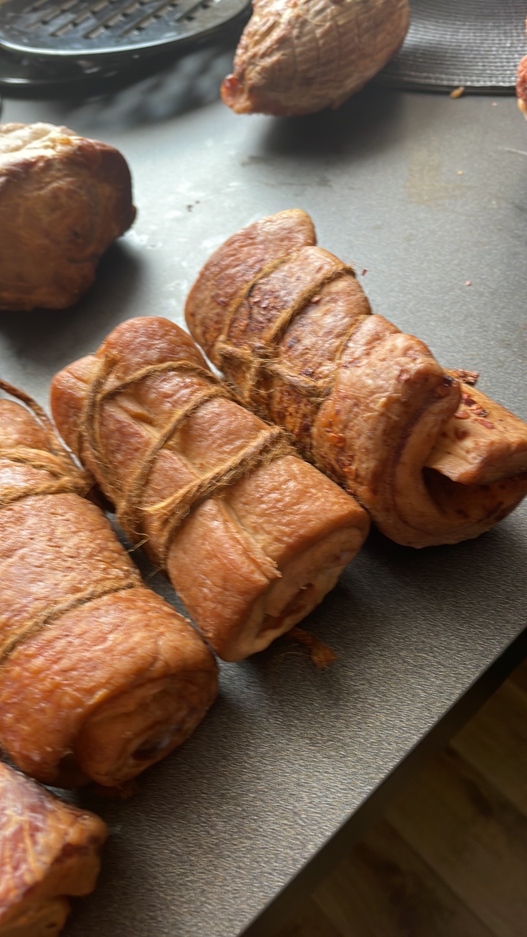 Wędliny wędzone, zawinięte sznurkiem, ułożone na szarym blacie, z widoczną fakturą powierzchni.
