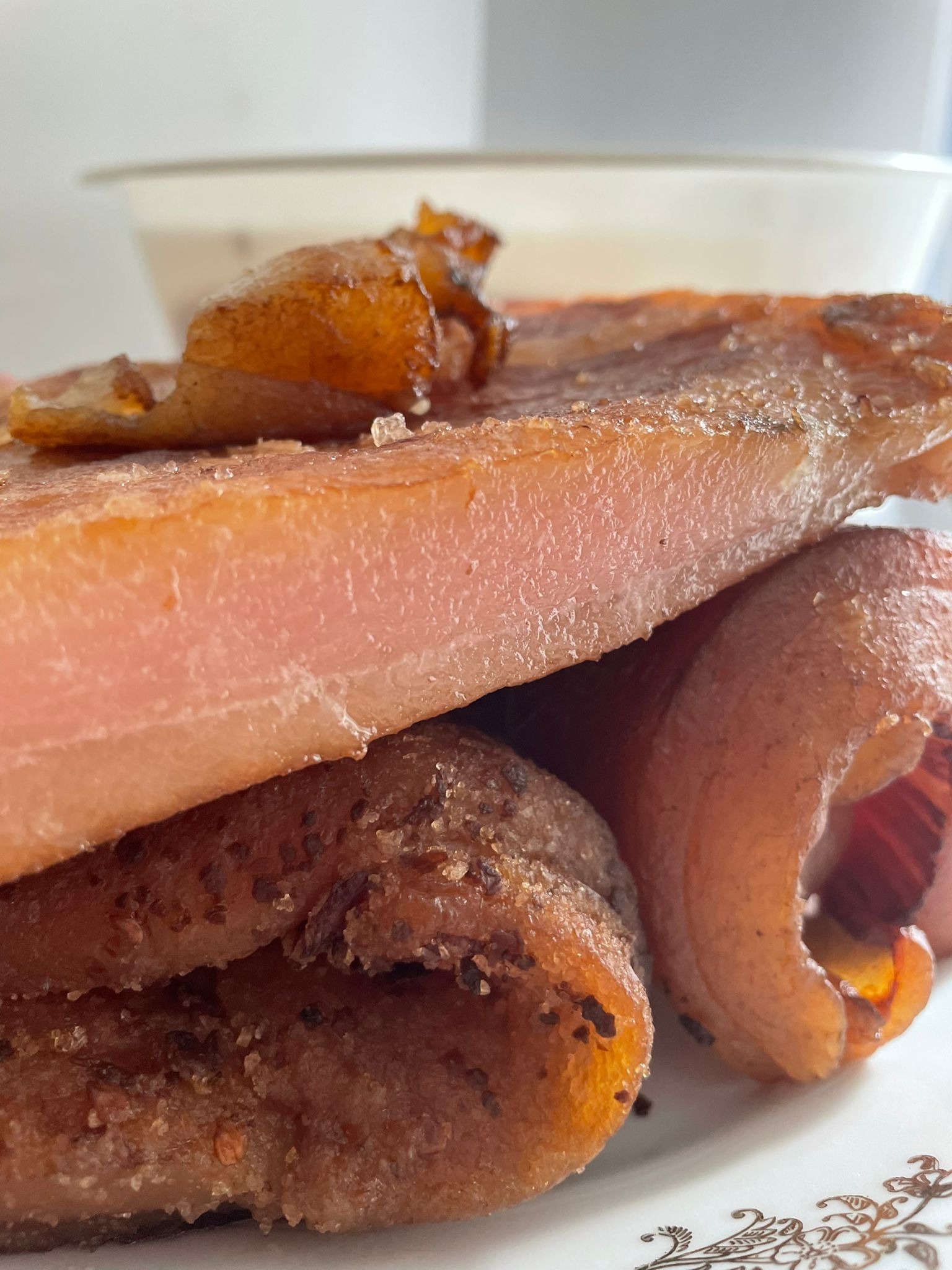 Zbliżenie na warstwową kompozycję z wędlin: plaster różowej szynki na wierzchu, pod nim smażone, brązowe krążki z przyprawami, całość na talerzu z delikatnym, złotym wzorem.