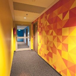 VIARTMEDIA MONIKA MINORCZYK - Korytarz z geometryczną grafiką ścienną w odcieniach żółci i pomarańczy, jasna żółta ściana po lewej, szara wykładzina podłogowa, drzwi ewakuacyjne na końcu korytarza.