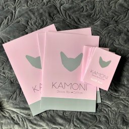 VIARTMEDIA MONIKA MINORCZYK - Stos różowych materiałów firmowych z logo w kształcie kociej głowy na tle szarej, wzorzystej tkaniny.