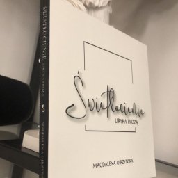 VIARTMEDIA MONIKA MINORCZYK - Biała okładka książki 'Światłocienie. Liryka prozą' autorstwa Magdaleny Ojrzyńskiej, ustawiona pionowo na półce, obok białej figury anioła.