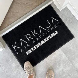 VIARTMEDIA MONIKA MINORCZYK - Czarna wycieraczka z białym logo 'KARKAJA Kaja Karpienko MAKEUP STUDIO' leżąca na szarej podłodze, widziane z góry, z fragmentem butów osoby stojącej na wycieraczce.