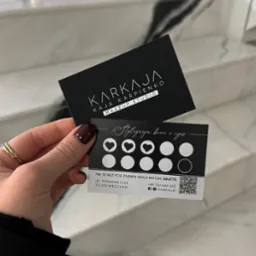 Dwie wizytówki studia makijażu Karkaja trzymane w dłoni na tle marmurowych schodów, jedna z logo, druga z ofertą stylizacji brwi i rzęs oraz informacją o darmowym zabiegu Gold Botox po 10 wizytach.