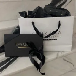 Elegancki zestaw prezentowy z logo firmy Karka Kaja Karpienko Makeup Artist we Wrocławiu, składający się z czarnego pudełka przewiązanego wstążką i białej torby z czarnymi uchwytami i logo,...