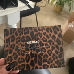 Karta z panterkowym wzorem marki KERMÉ promująca kredkę do ust Day & Night Lip Liner trzymana w dłoni z czarnym manicure, w tle torba prezentowa i roślina doniczkowa.