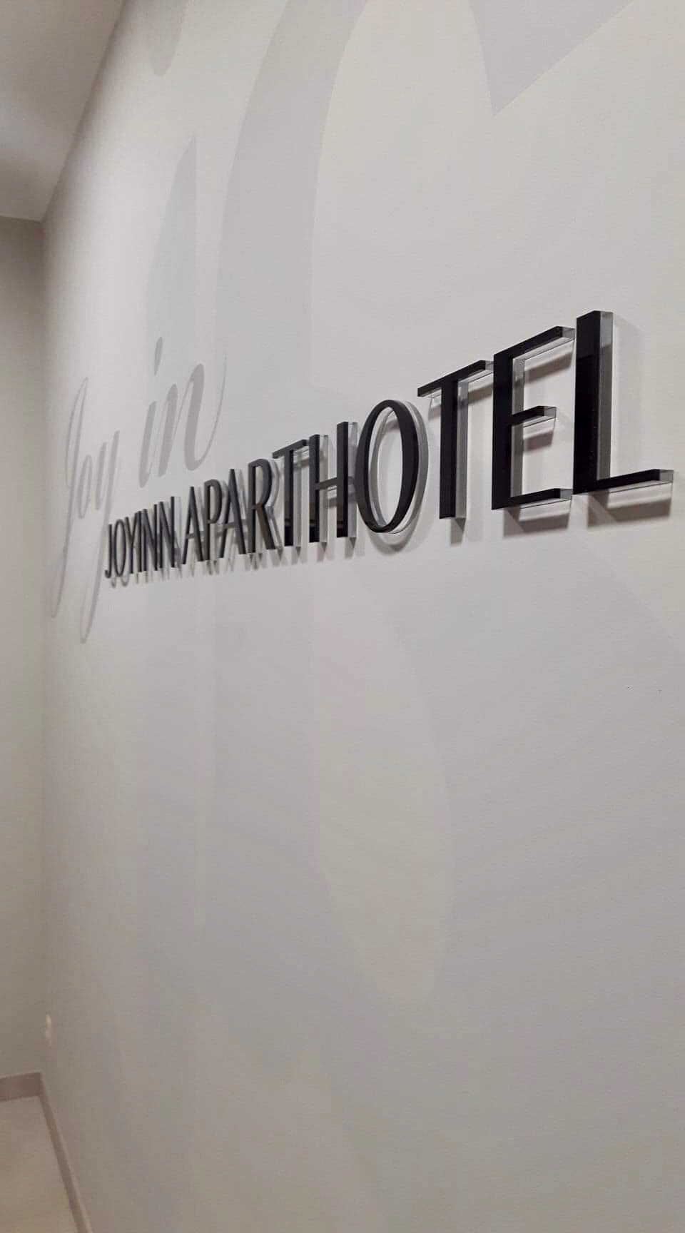 Ścienne logo 'Joyinn Aparthotel' wykonane z czarnych, trójwymiarowych liter z efektem cienia, umieszczone na białej ścianie z delikatnym, jasnym napisem w tle.