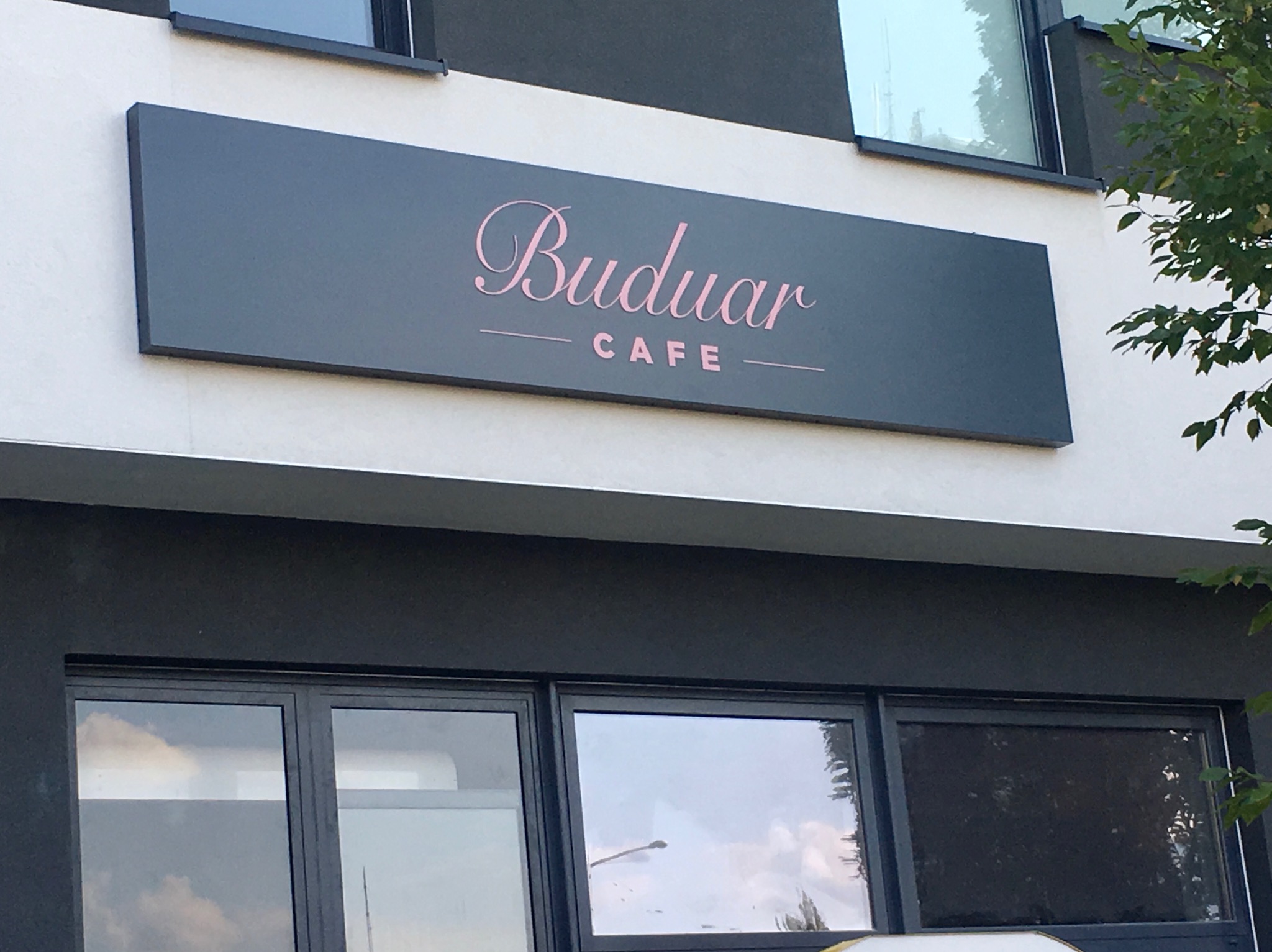 Szyld kawiarni Buduar Cafe z różowym napisem, umieszczony na białej fasadzie budynku z ciemnymi oknami.