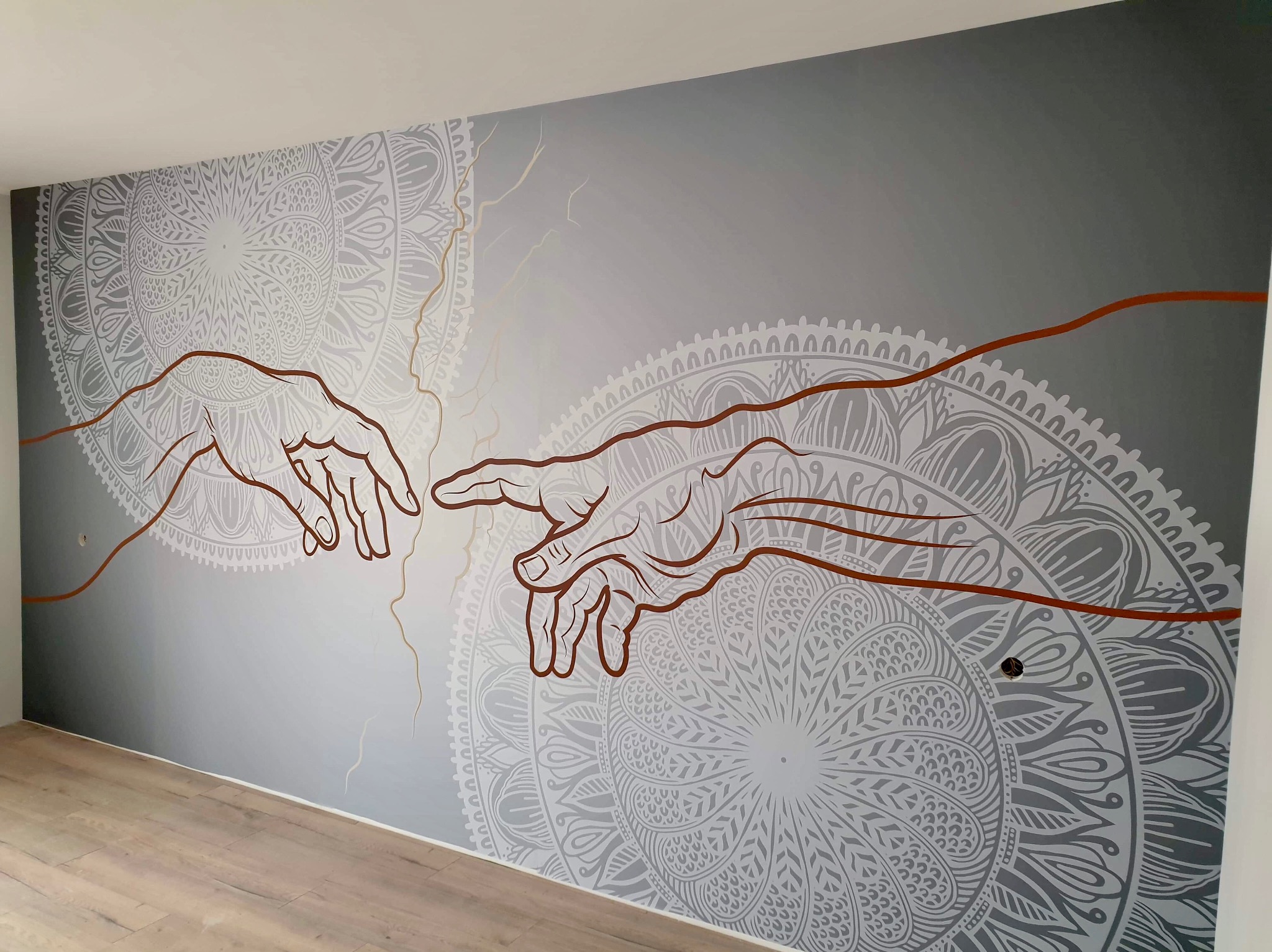 Szary mural na ścianie przedstawiający dłonie inspirowane 'Stworzeniem Adama' na tle mandali, wykonany farbami w odcieniach szarości i brązu w minimalistycznym wnętrzu.