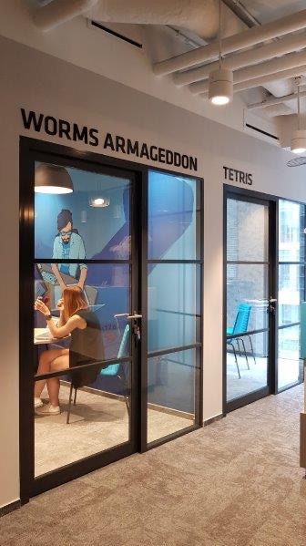 Korytarz biurowy z przeszklonymi salami o nazwach gier komputerowych: Worms Armageddon z kobietą siedzącą przy biurku i Tetris z widocznymi krzesłami.