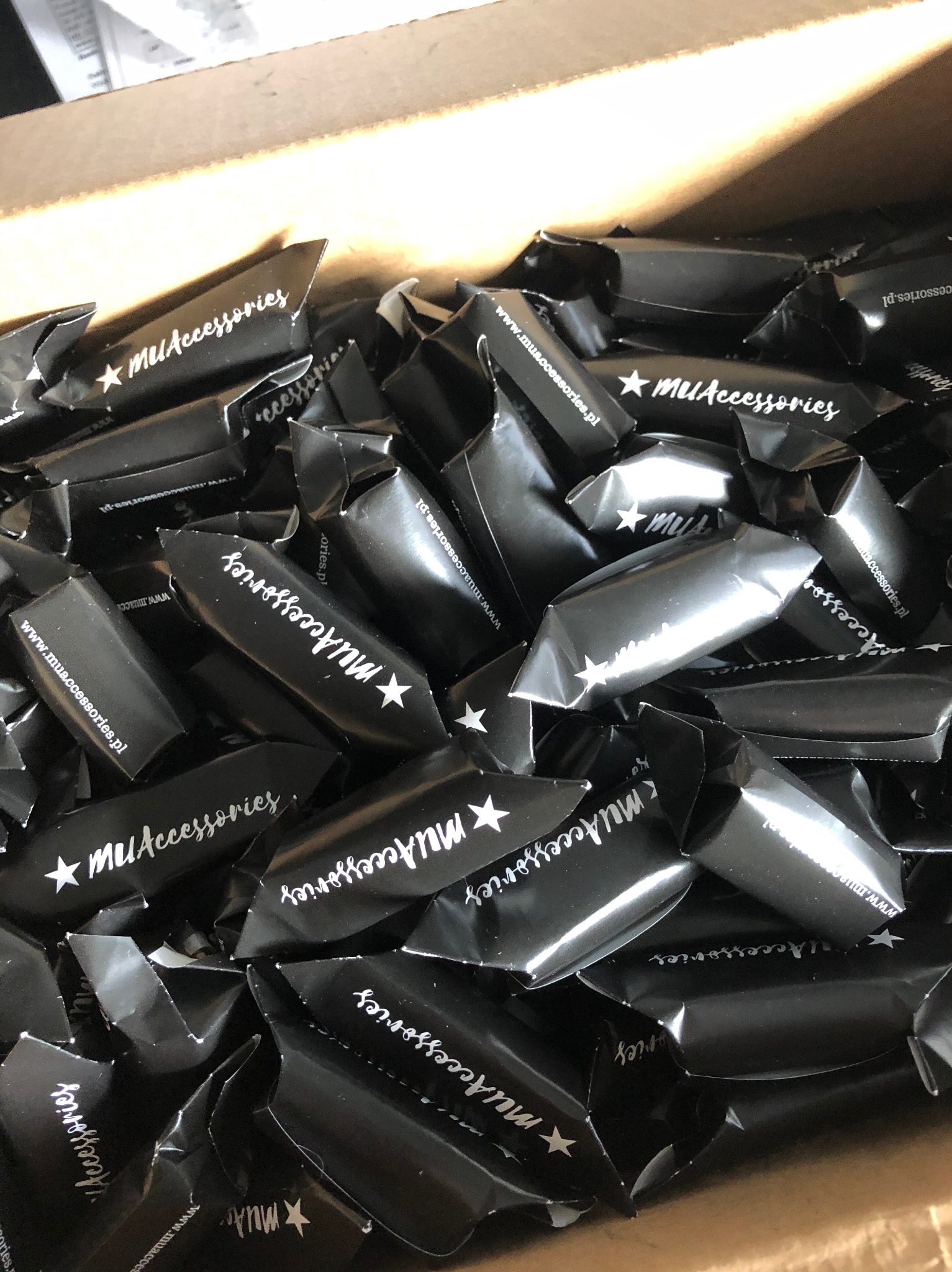 Stos czarnych, błyszczących opakowań cukierków z białym nadrukiem logo MUAccessories i adresem strony internetowej, ułożonych w kartonowym pudełku.