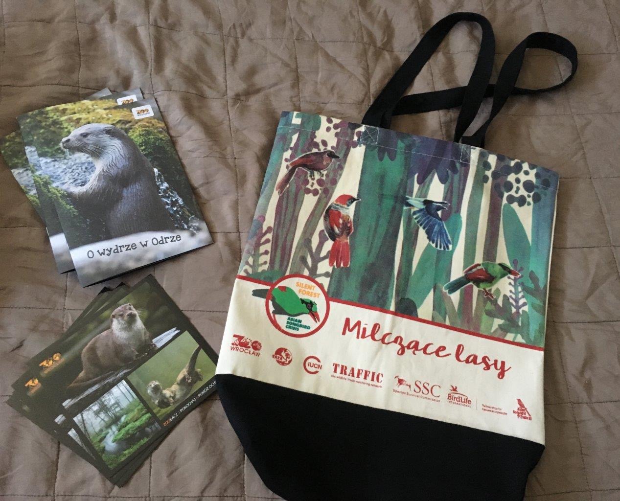 Bawełniana torba z nadrukiem kolorowych ptaków i napisem 'Milczące lasy' leży obok stosu pocztówek z wizerunkiem wydry na brązowym tle.