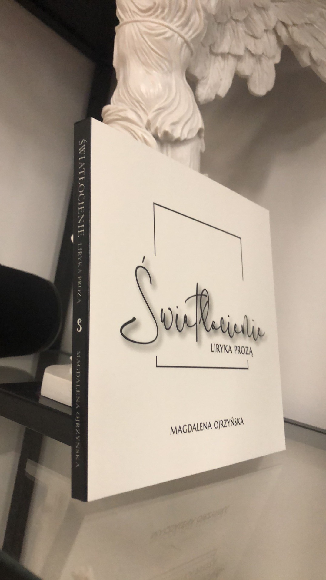 Biała okładka książki 'Światłocienie. Liryka prozą' autorstwa Magdaleny Ojrzyńskiej, ustawiona pionowo na półce, obok białej figury anioła.