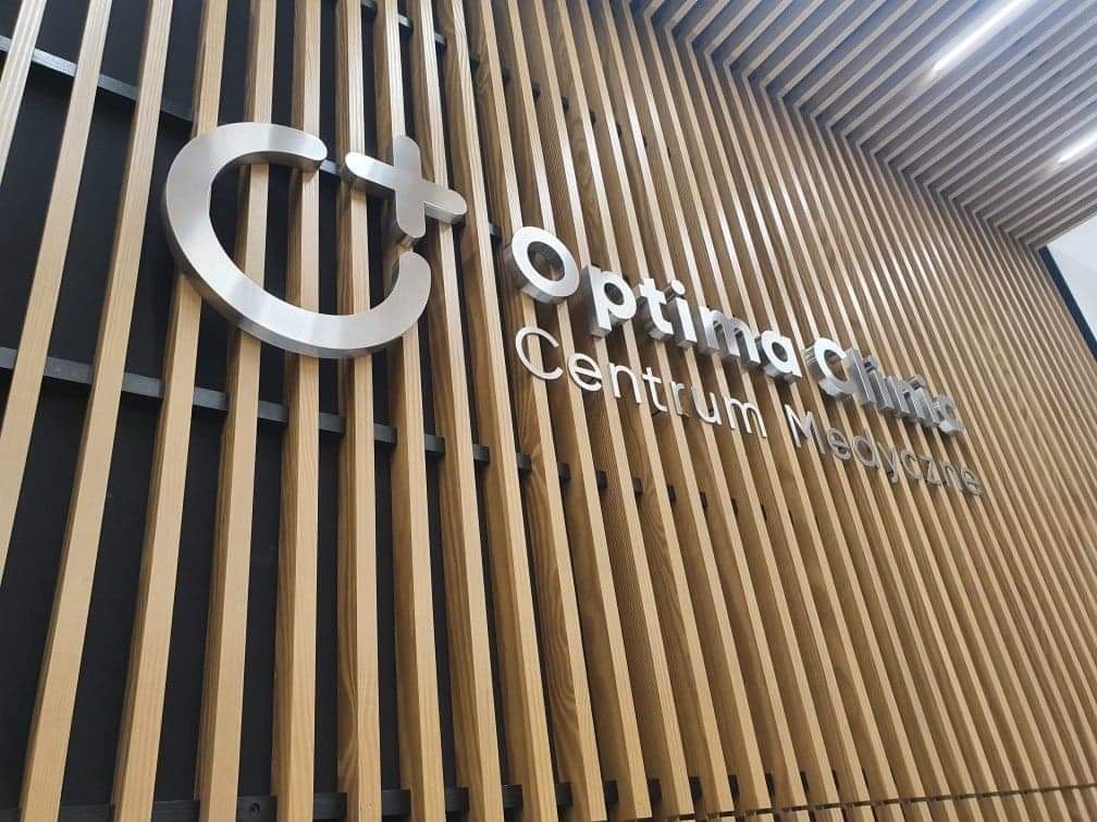 Metalowe logo 'C+' i napis 'Optima Clinic Centrum Medyczne' na drewnianej ścianie z pionowych listew, widok z dołu.