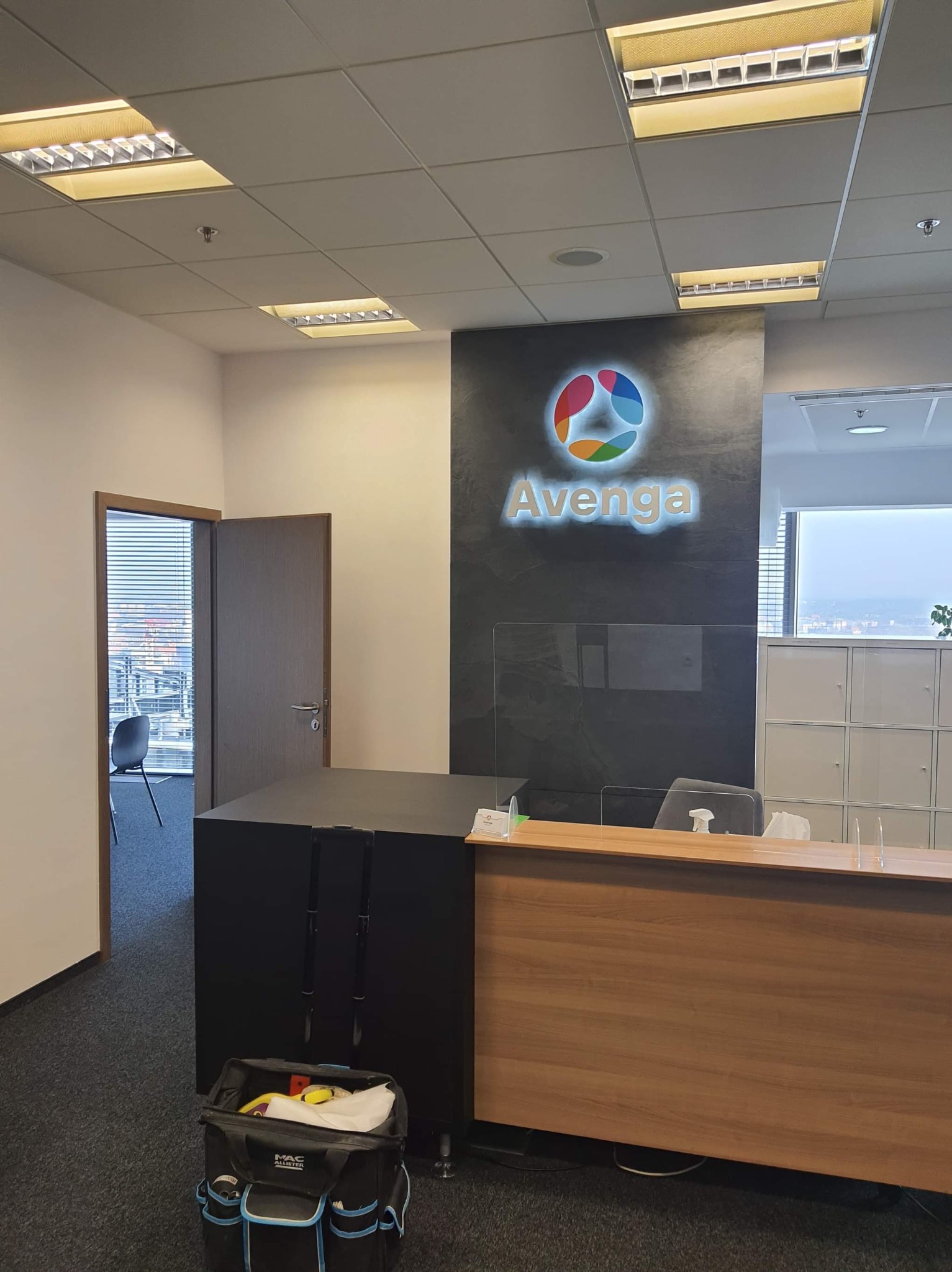 Recepcja biura z podświetlanym logo Avenga na czarnej ścianie, widoczny blat recepcji z pleksi, narzędzia w torbie na podłodze, otwarte drzwi do biura z widokiem na miasto przez żaluzje.