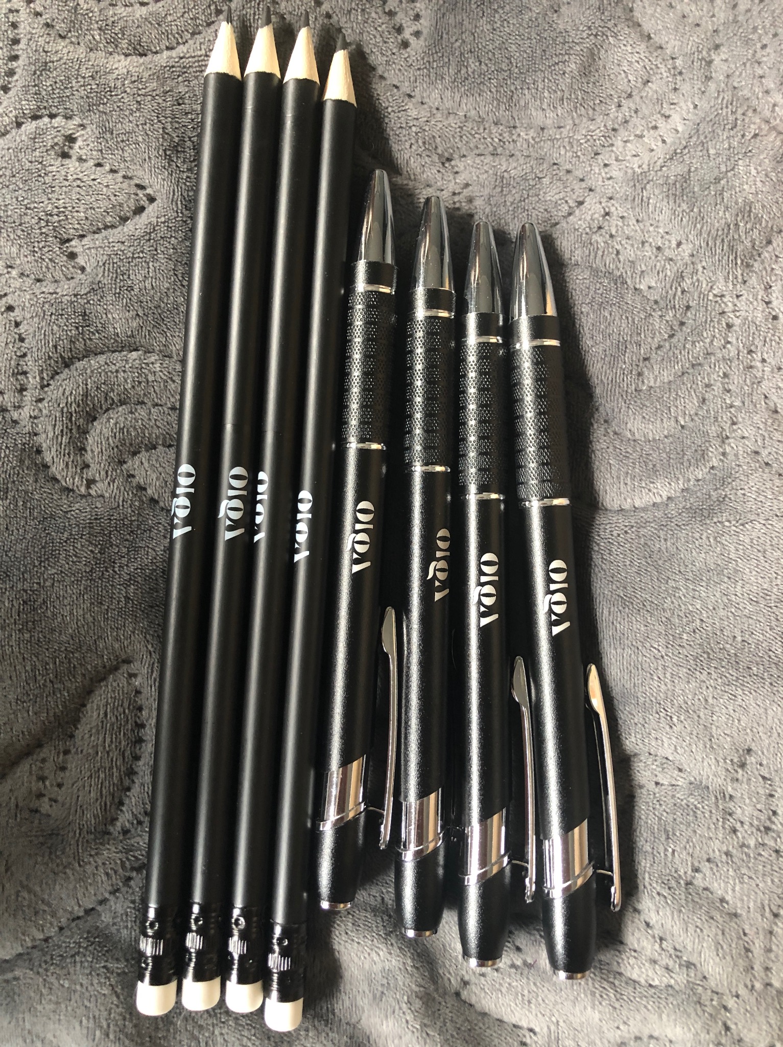Cztery czarne ołówki i cztery czarne długopisy z srebrnymi elementami i białym napisem 'VOIO' ułożone na szarym, teksturowanym tle.