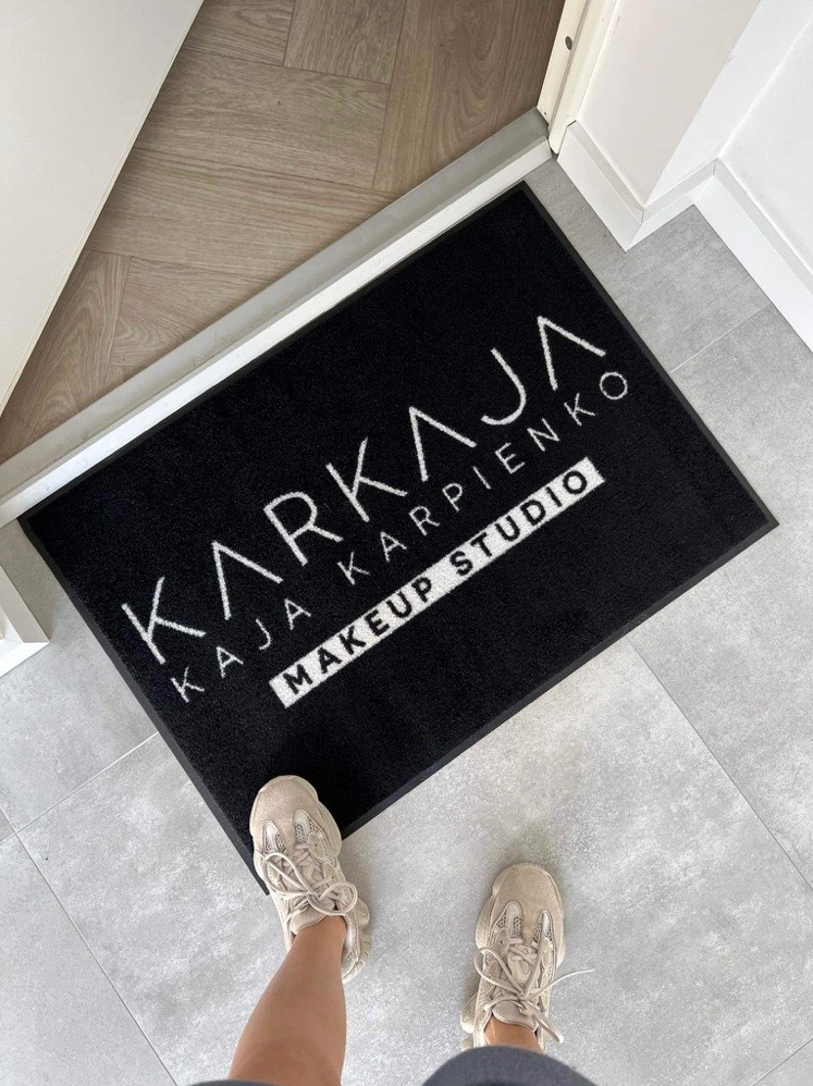 Czarna wycieraczka z białym logo 'KARKAJA Kaja Karpienko MAKEUP STUDIO' leżąca na szarej podłodze, widziane z góry, z fragmentem butów osoby stojącej na wycieraczce.