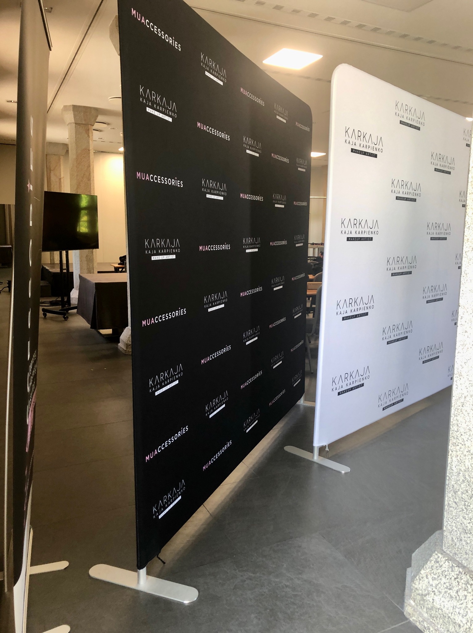 Dwa ścianki reklamowe z nadrukowanymi logotypami i nazwami 'KARKAJA KAJA KARPIENKO MAKE UP ARTIST' oraz 'MUACCESSORIES', stojące w jasnym wnętrzu z kolumnami i ciemnymi podłogami.