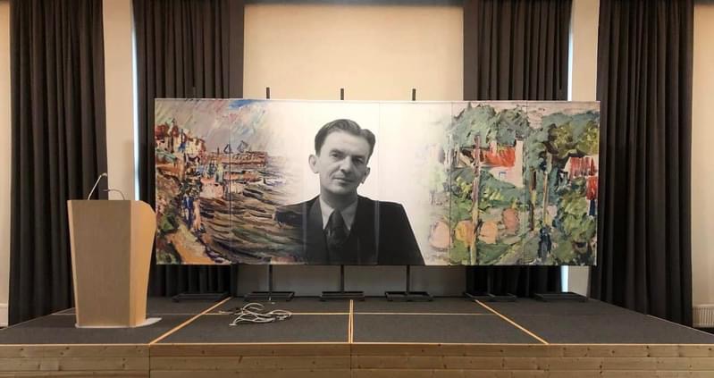 Scena z mównicą i dużym banerem przedstawiającym czarno-biały portret mężczyzny na tle abstrakcyjnego pejzażu miejskiego i wiejskiego.