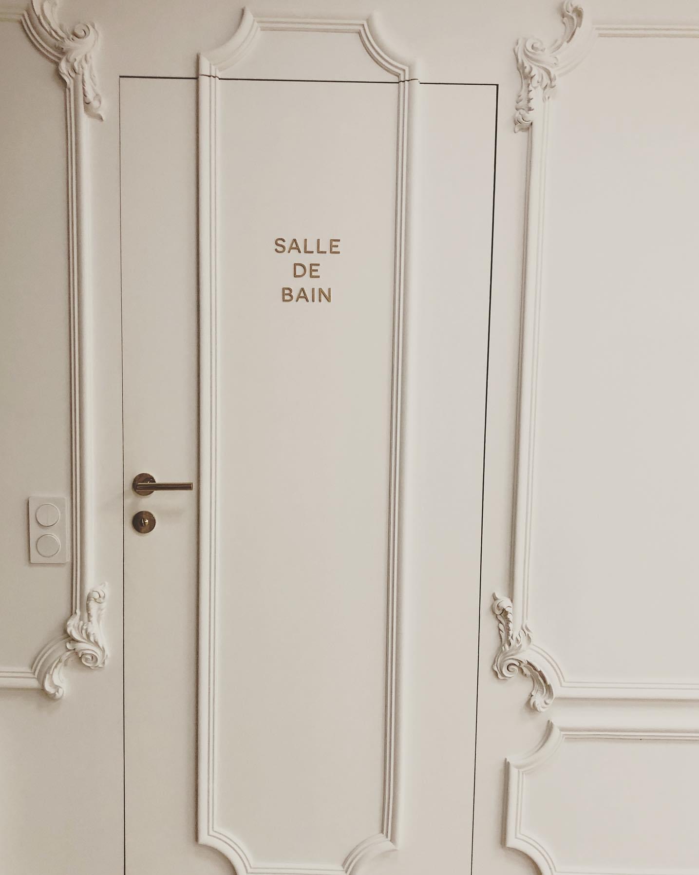 Białe drzwi do łazienki z ozdobnymi sztukateriami i złotym napisem 'SALLE DE BAIN', widoczny włącznik światła i złota klamka.