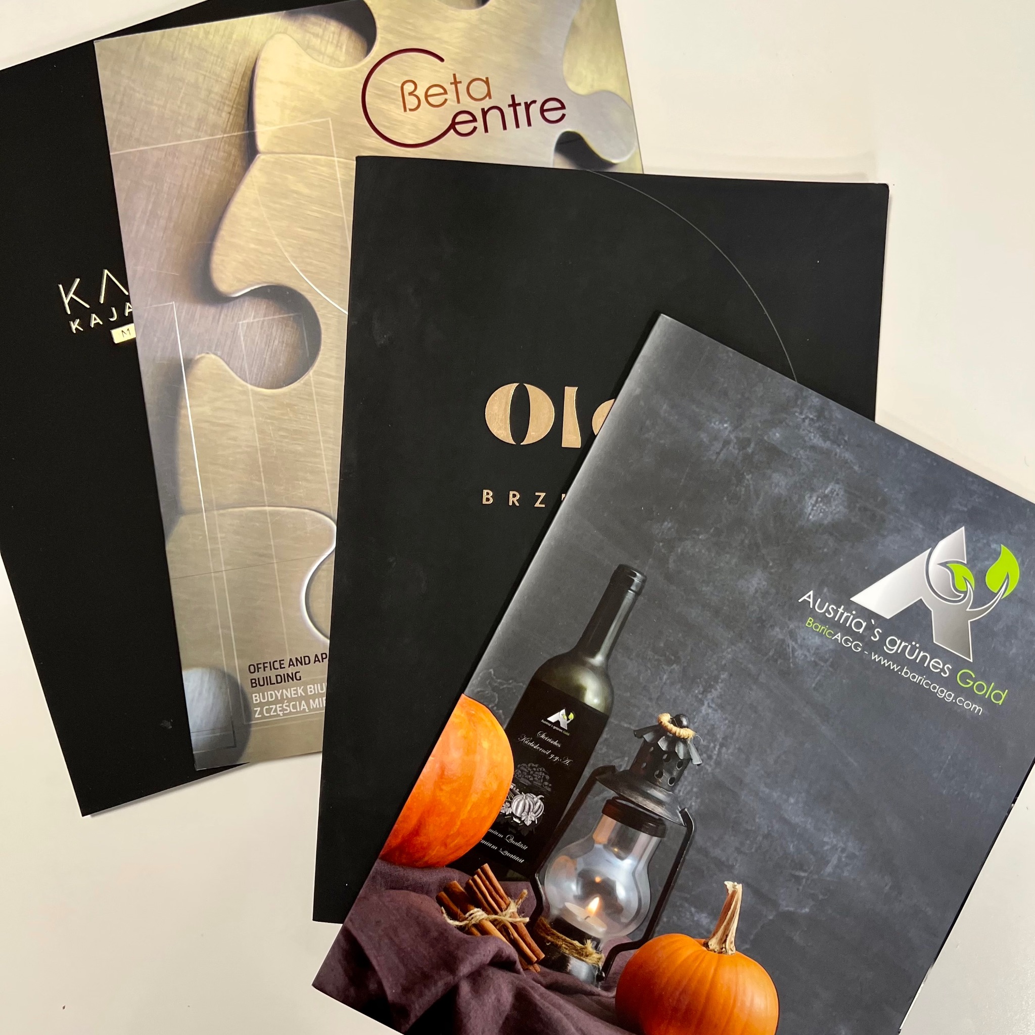 Stos czterech folderów reklamowych z różnymi projektami graficznymi, w tym folder z motywem jesiennym z dyniami i lampionem.