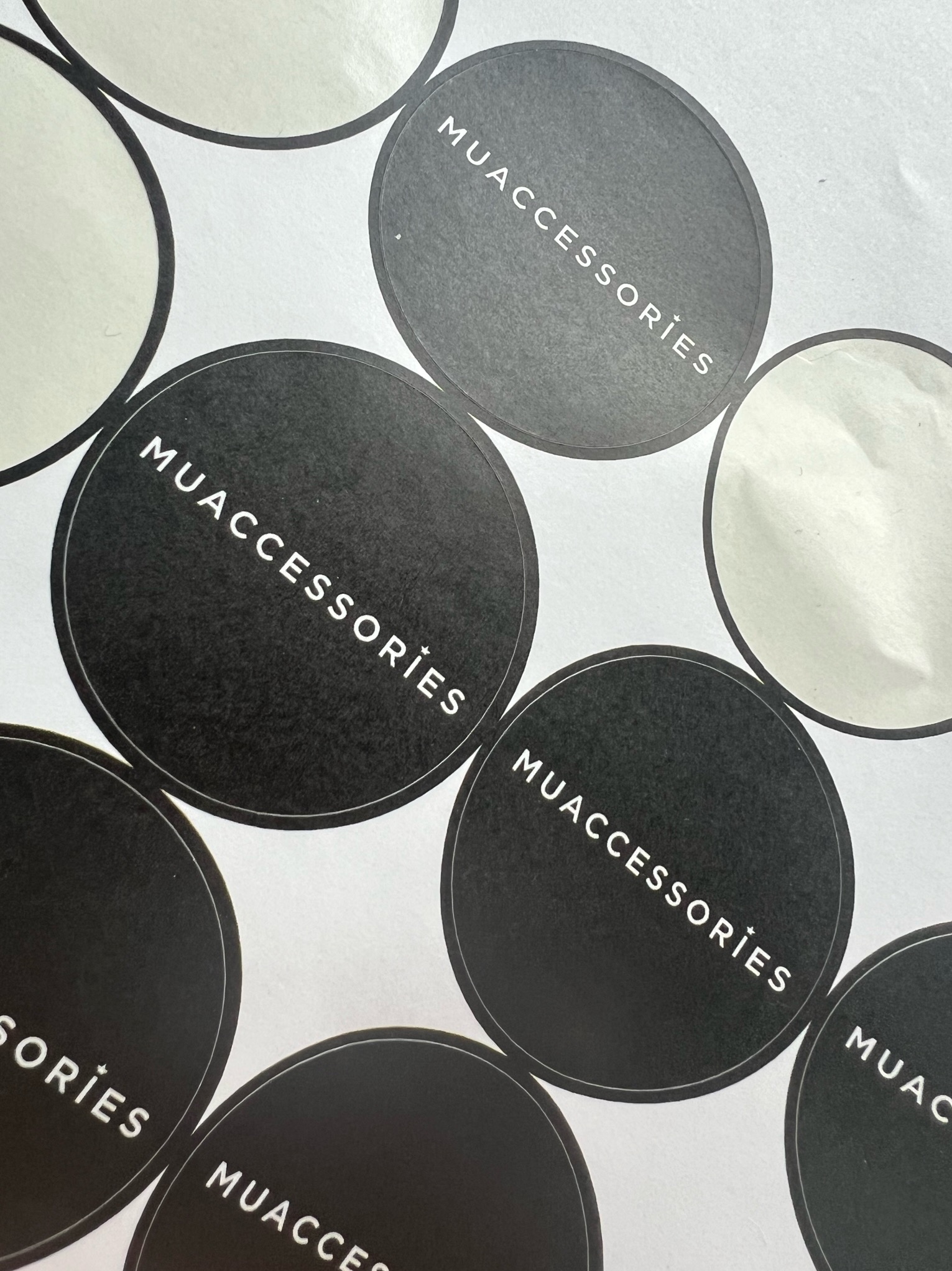 Arkusz z okrągłymi, czarnymi naklejkami z białym napisem 'MUACCESSORIES' w ukośnym układzie.