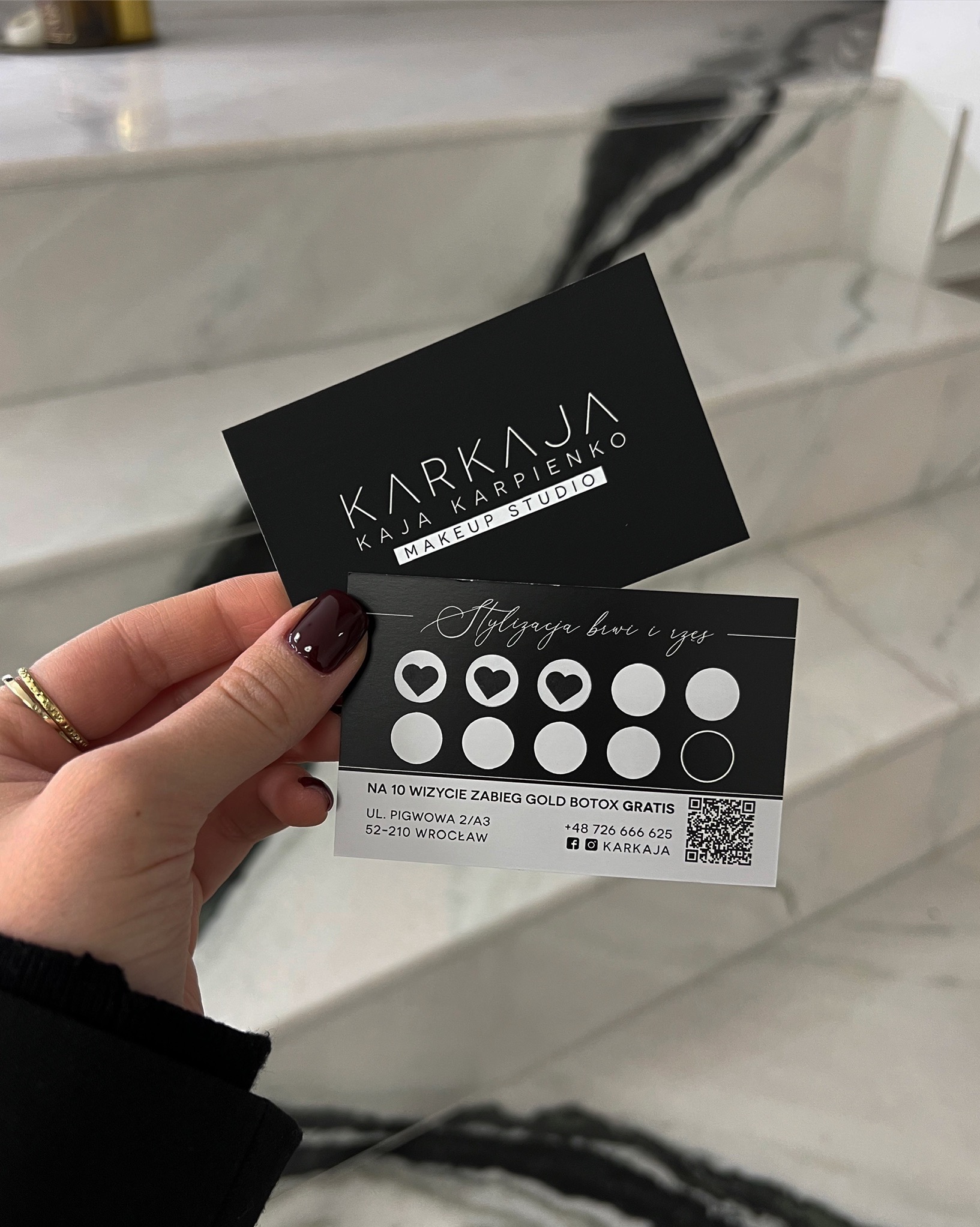 Dwie wizytówki studia makijażu Karkaja trzymane w dłoni na tle marmurowych schodów, jedna z logo, druga z ofertą stylizacji brwi i rzęs oraz informacją o darmowym zabiegu Gold Botox po 10 wizytach.