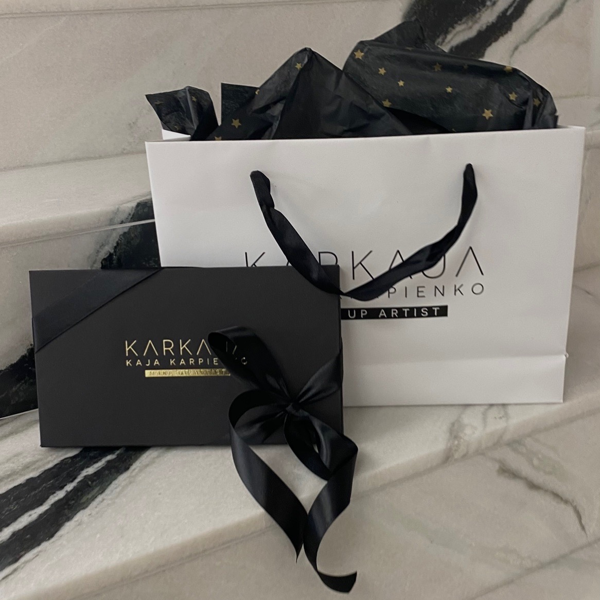 Elegancki zestaw prezentowy z logo firmy Karka Kaja Karpienko Makeup Artist we Wrocławiu, składający się z czarnego pudełka przewiązanego wstążką i białej torby z czarnymi uchwytami i logo,...