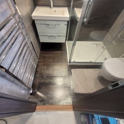 High standard Wykończeniówka - Nowoczesna łazienka z kabiną prysznicową, umywalką z szafką, grzejnikiem drabinkowym i podłogą imitującą drewno, widziana z perspektywy osoby stojącej w progu, z widocznymi butami i fragmentem...