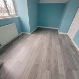 Jasnoszara podłoga z paneli ułożona w pokoju z błękitnymi ścianami i białymi listwami przypodłogowymi, widok na okno z kratownicą.