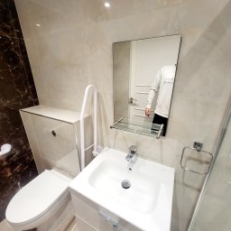 High standard Wykończeniówka - Nowoczesna, jasna łazienka z umywalką na szafce, toaletą z ukrytą spłuczką i uchwytem dla osób z niepełnosprawnościami, wykończona płytkami imitującymi marmur, z odbiciem osoby w lustrze.