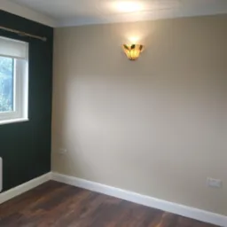 Pokój po remoncie z ciemnozieloną ścianą, panelami podłogowymi w odcieniach brązu i beżową ścianą z ozdobnym kinkietem.