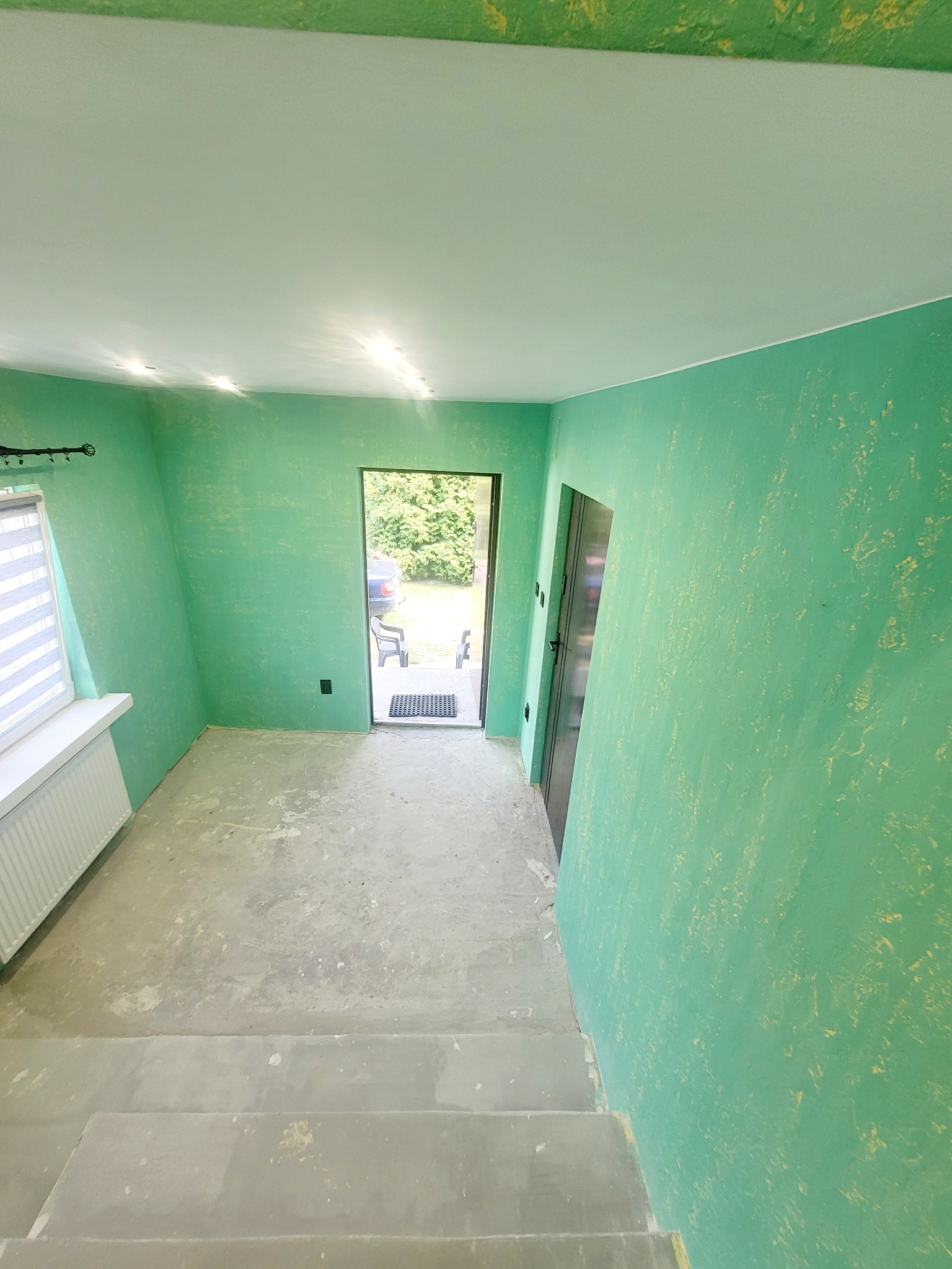 Wnętrze w trakcie remontu z zielonymi ścianami o nieregularnej fakturze, surowy betonowy podkład podłogowy, schody i widok na zewnątrz przez otwarte drzwi.