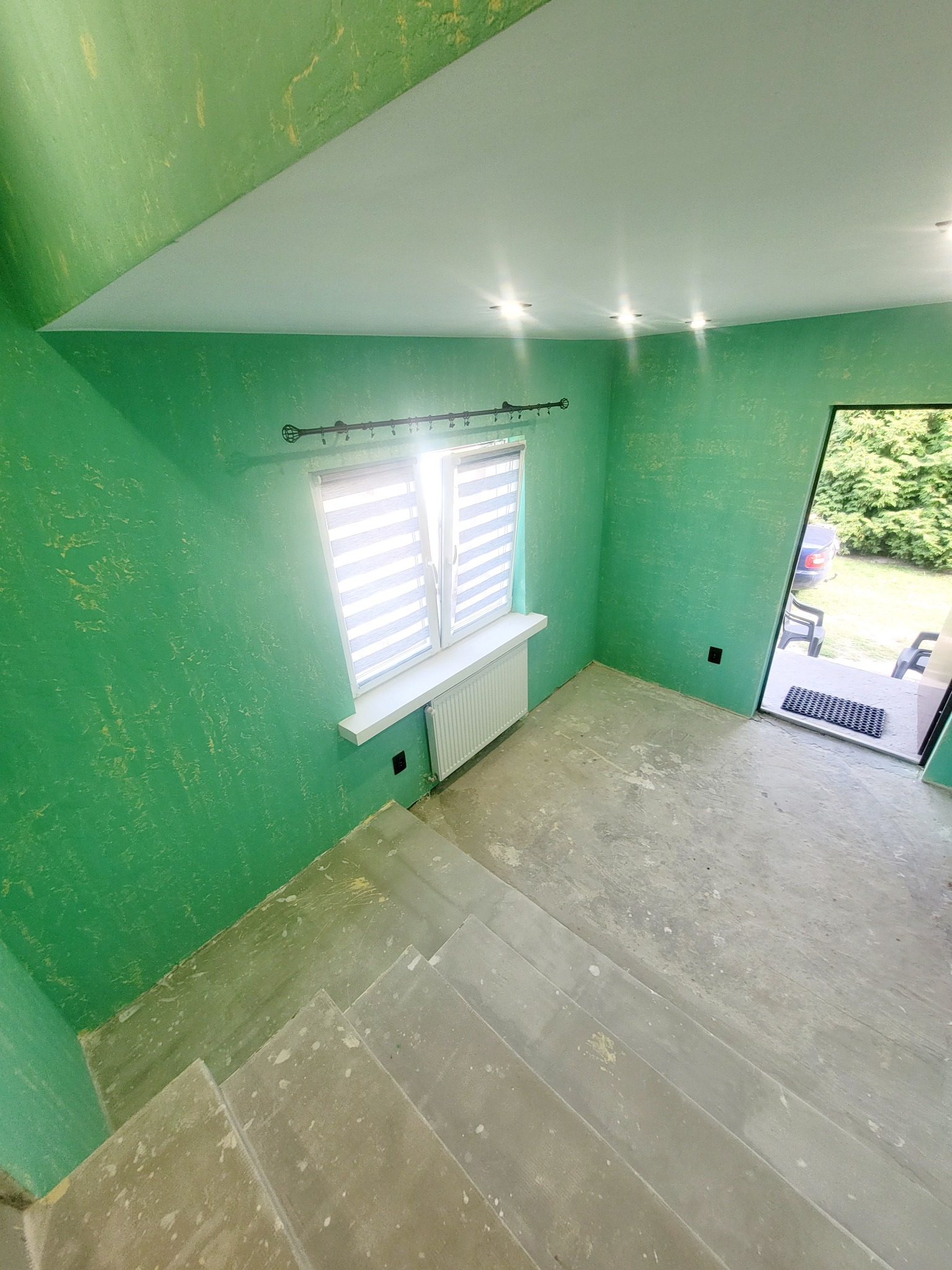 Wnętrze w trakcie prac wykończeniowych: ściany pomalowane na zielono z efektem dekoracyjnym, okno z żaluzjami, surowa podłoga ze schodami i wyjście na balkon z widokiem na zieleń.