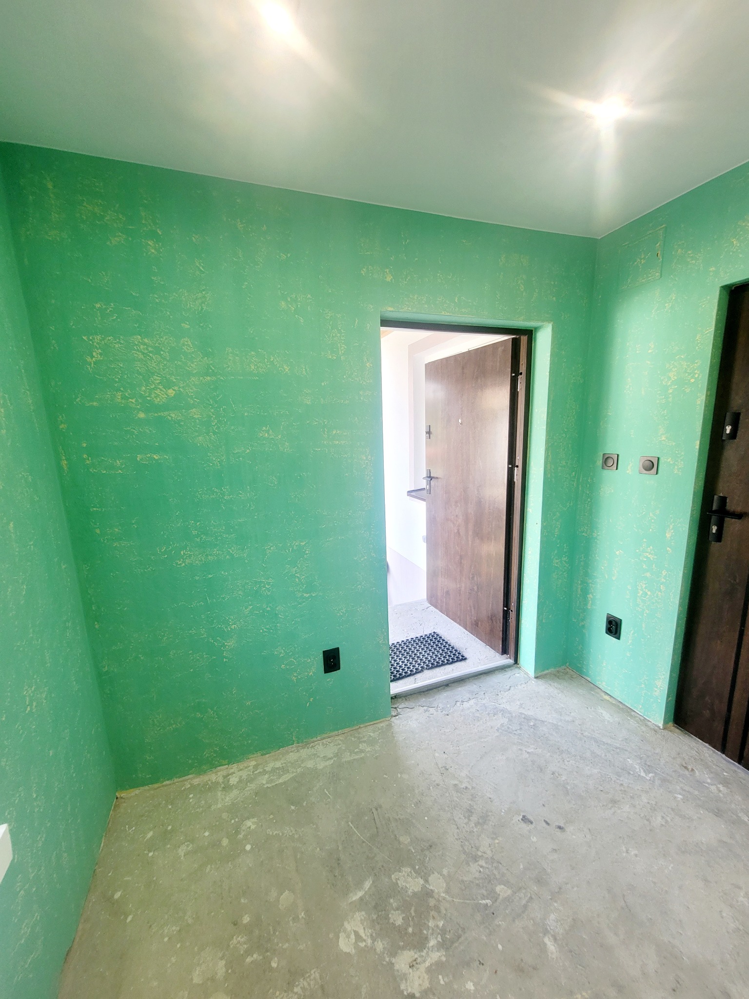 Wnętrze pokoju z zielonymi ścianami o nieregularnej fakturze, surową betonową podłogą i widocznymi instalacjami elektrycznymi, z otwartymi drzwiami wejściowymi w tle.