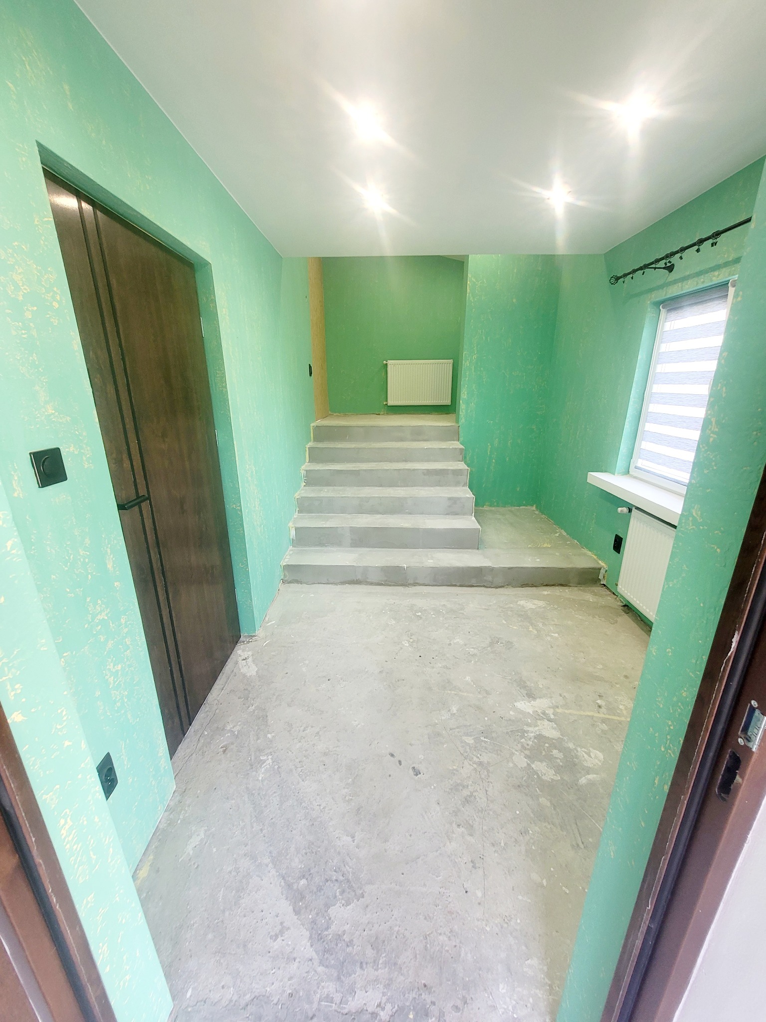 Wnętrze w trakcie prac wykończeniowych: zielone ściany z fakturą, surowe betonowe schody, widoczne elementy instalacji grzewczej i okno z żaluzjami. Otwory drzwiowe po bokach kadru.