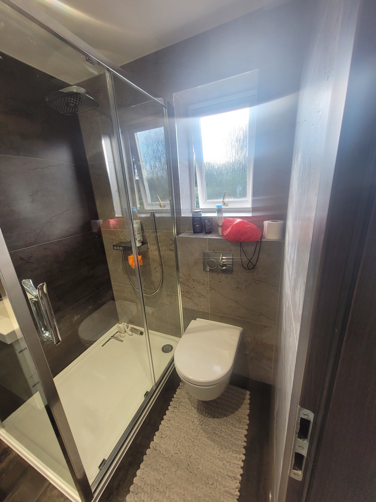 Nowoczesna, wąska łazienka z kabiną prysznicową typu walk-in, podwieszaną toaletą, oknem z widokiem na zieleń i dywanikiem z grubym włosiem.