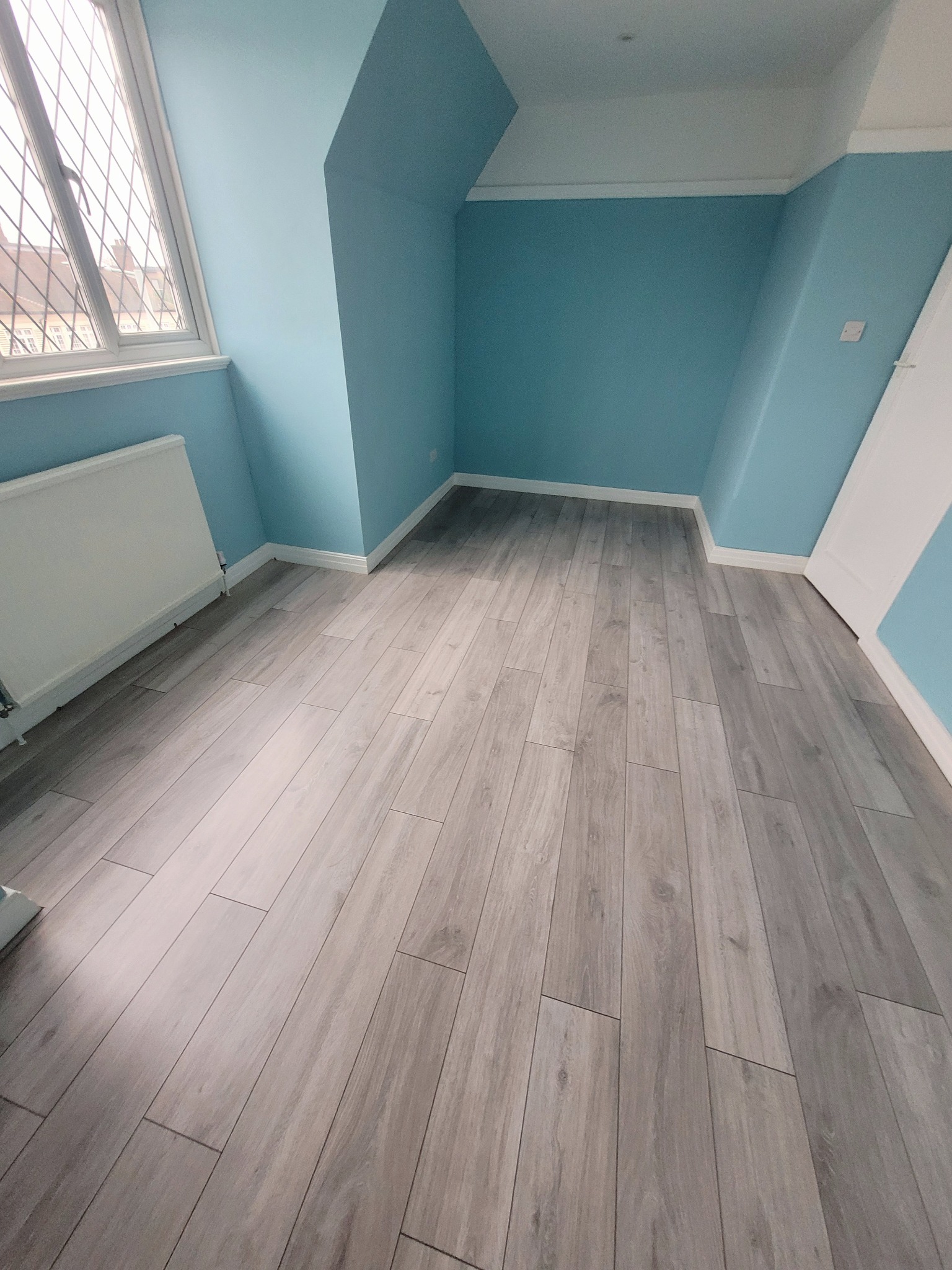 Jasnoszara podłoga z paneli ułożona w pokoju z błękitnymi ścianami i białymi listwami przypodłogowymi, widok na okno z kratownicą.