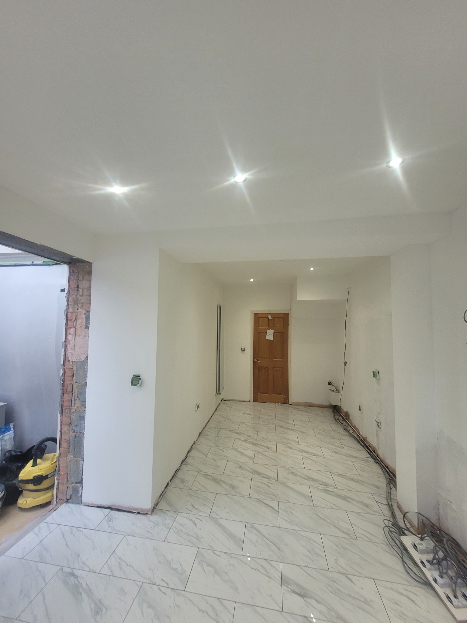 Wnętrze po remoncie z ułożonymi białymi płytkami imitującymi marmur, świeżo pomalowanymi ścianami i sufitem z zamontowanymi punktowymi lampami. Widoczne gniazdka elektryczne, przewody...