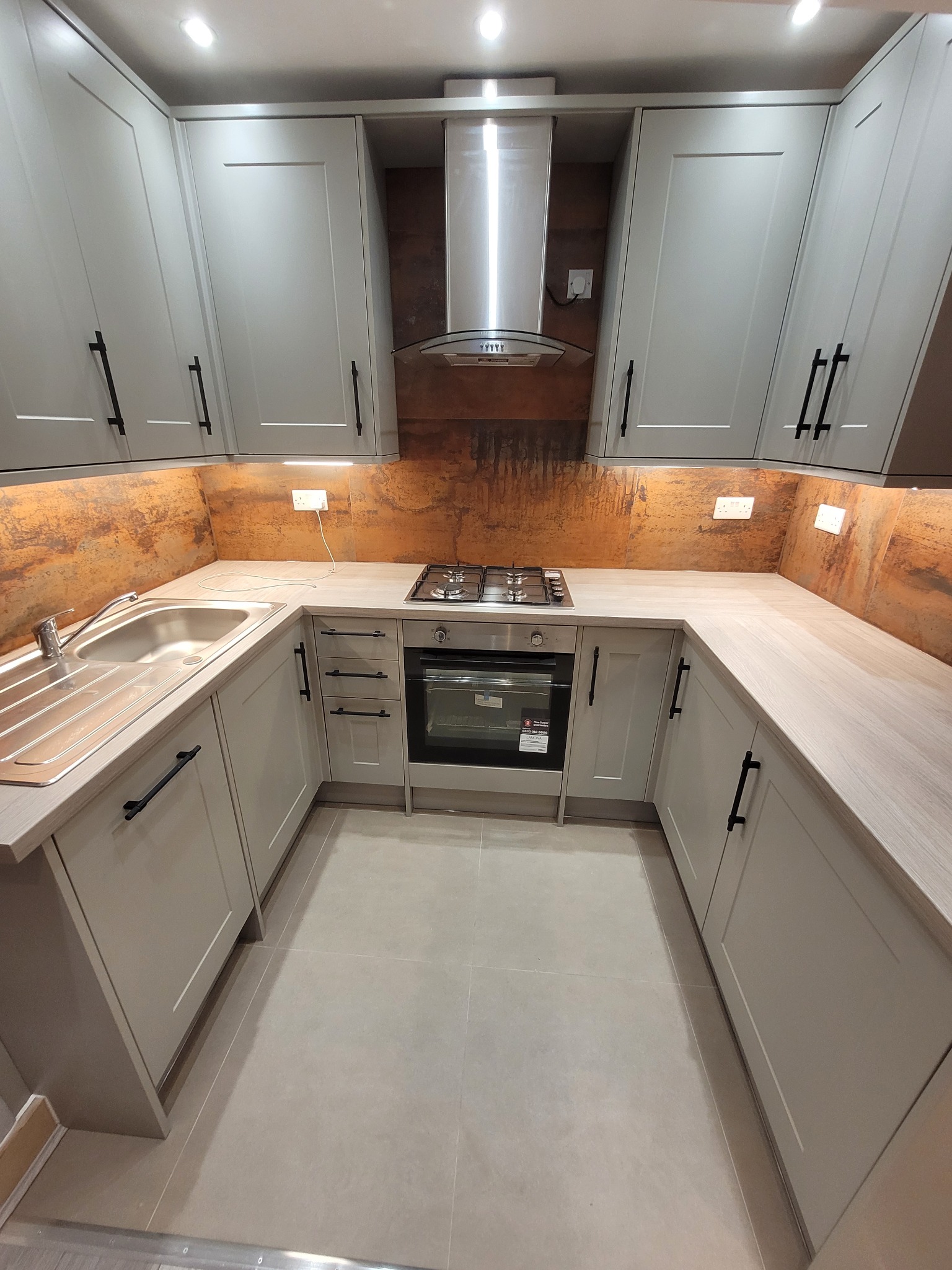 Kompaktowa kuchnia w kształcie litery U z szarymi szafkami, czarnymi uchwytami i blatem imitującym drewno, oświetlona ciepłym światłem pod szafkami wiszącymi, z rdzawym panelem nad blatem i płytą...