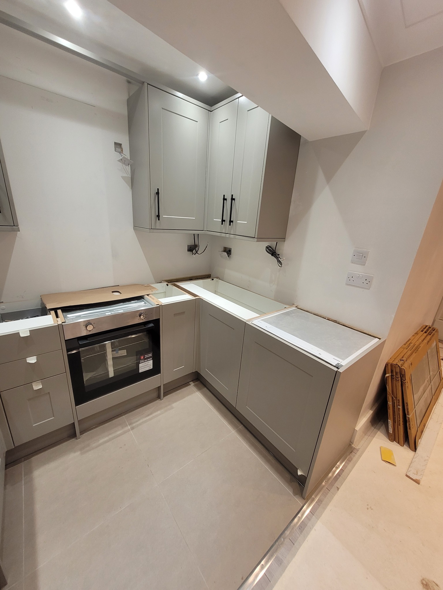 Montaż szarej kuchni z czarnymi uchwytami, widoczny piekarnik, brakujące blaty, kartony z panelami przy ścianie, gniazdka elektryczne na ścianie, stan surowy.