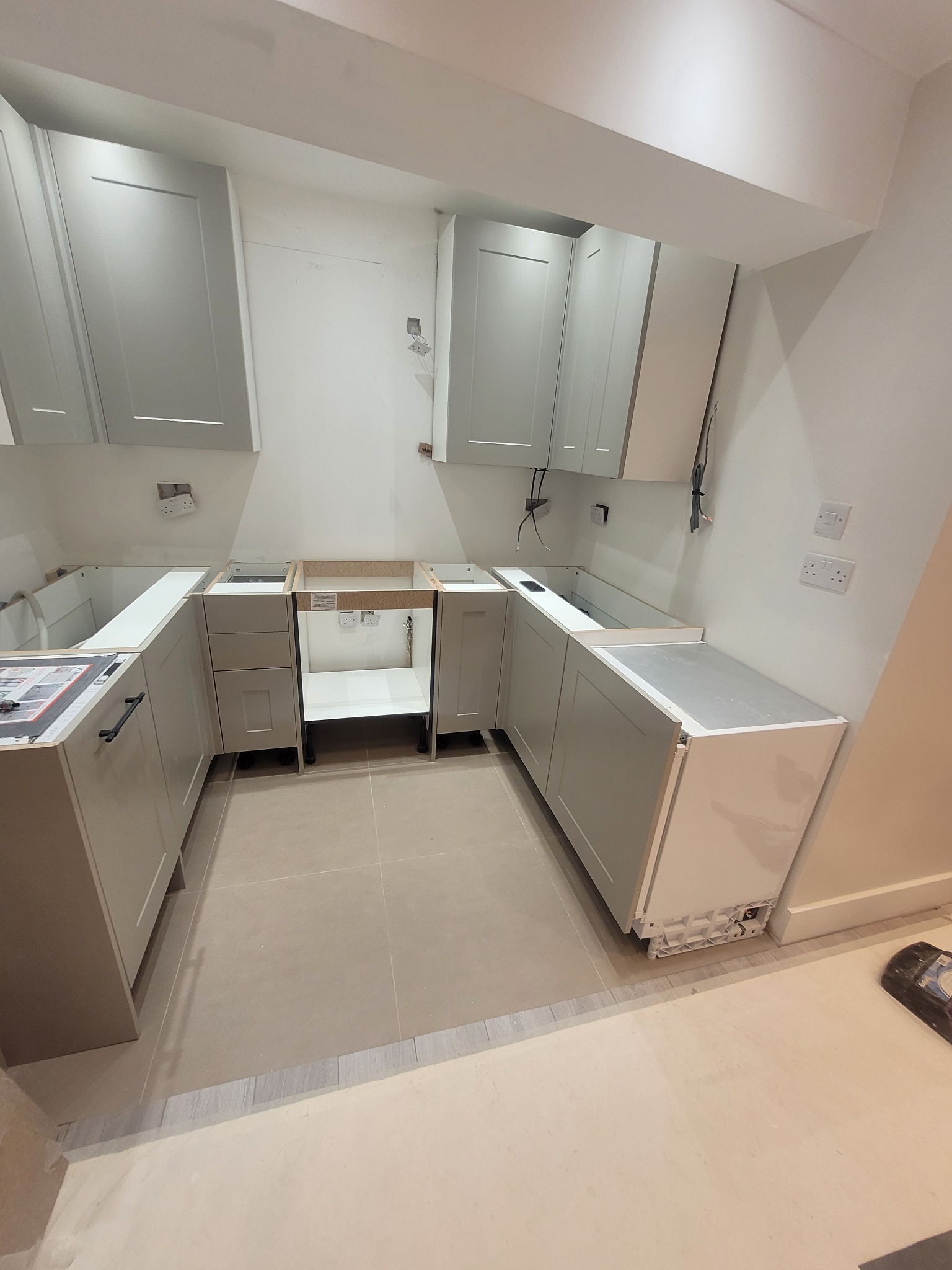 Montaż szarych szafek kuchennych, widoczne wiszące szafki i dolne moduły bez blatów, na podłodze jasne płytki, widoczne gniazdka elektryczne na ścianie.