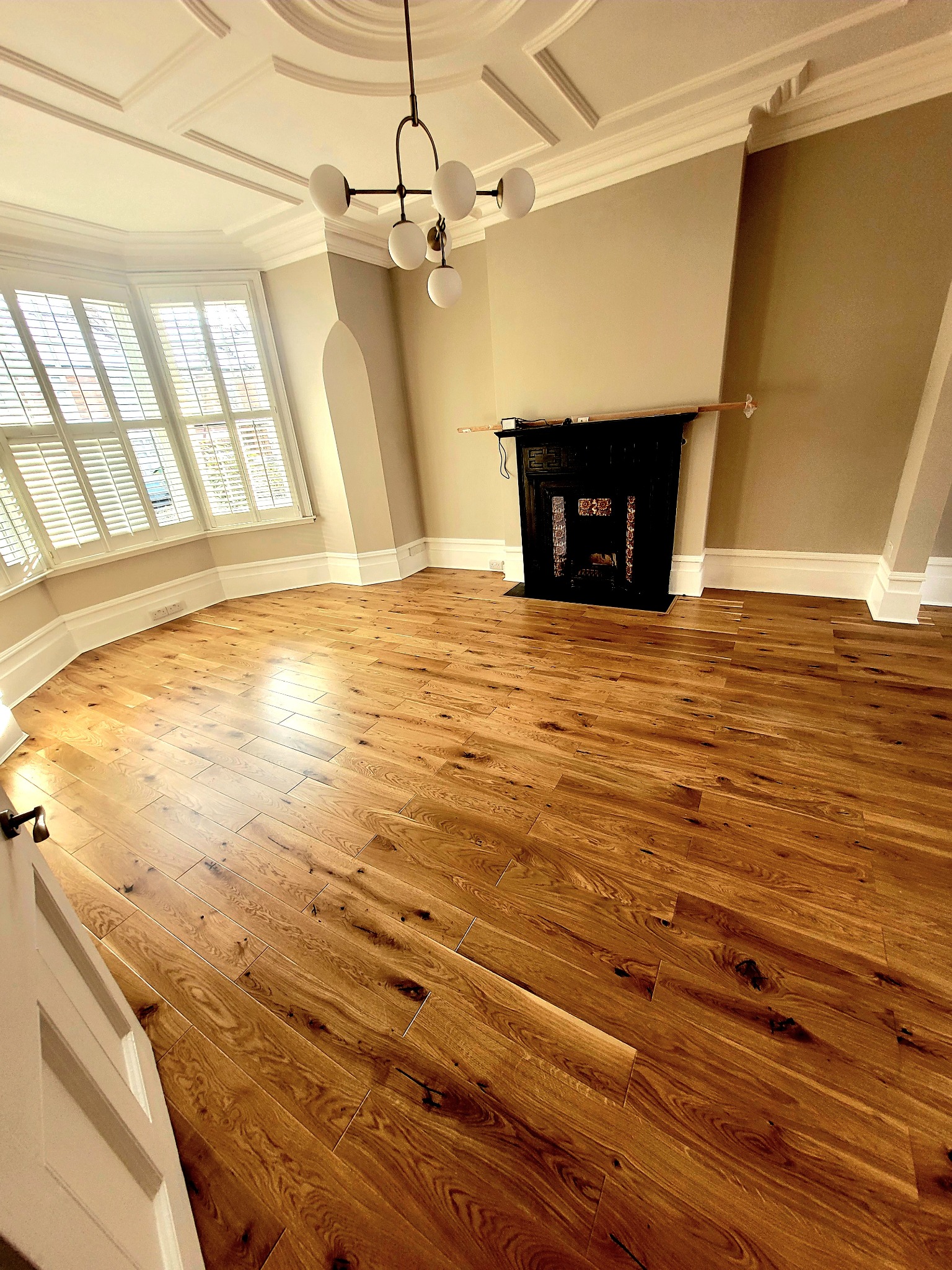 Wykończona podłoga z naturalnego drewna w salonie w Łęcznej, widok na kominek i okno z żaluzjami, nowoczesna lampa wisząca.