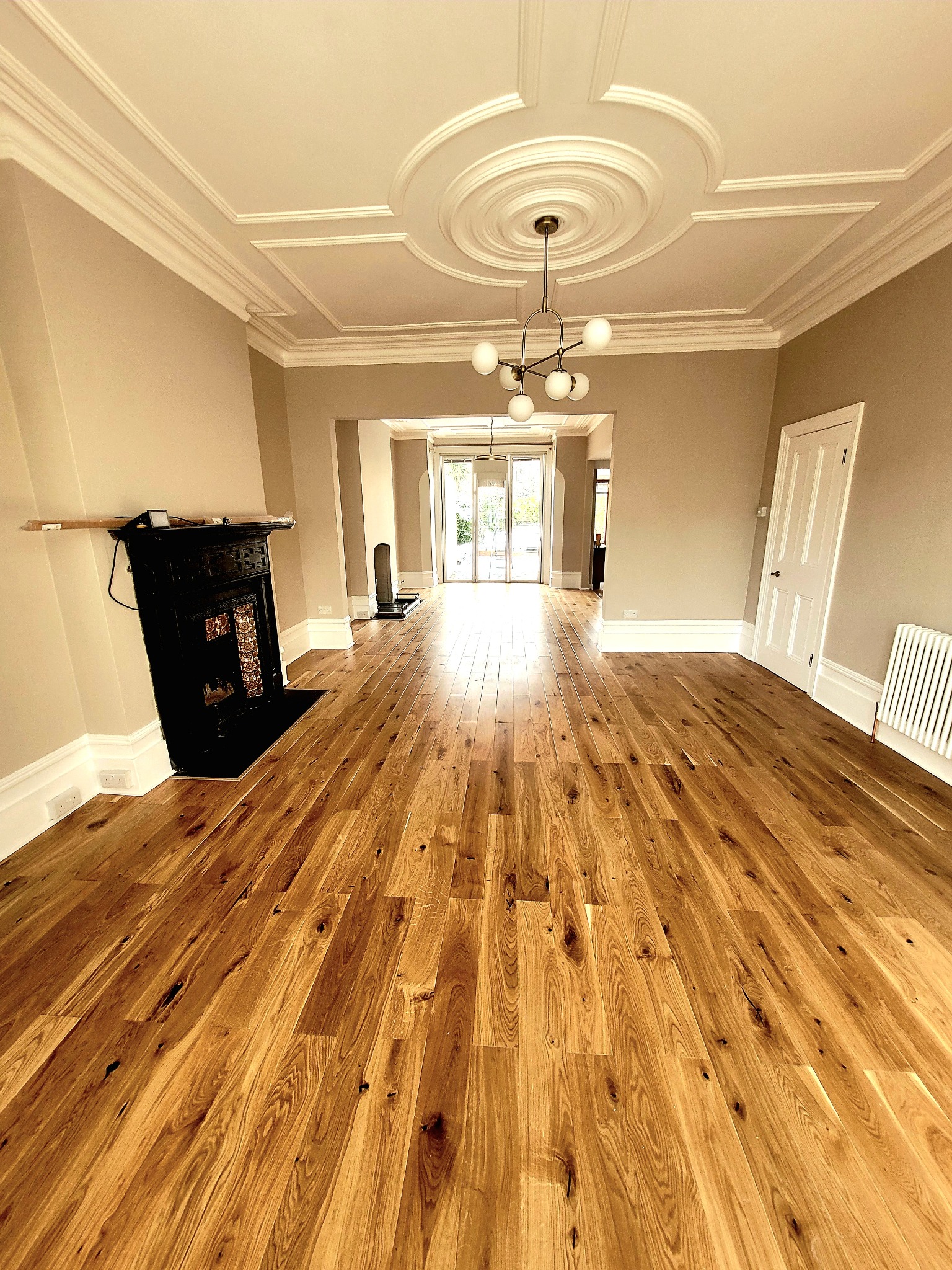 Salon z nową, jasną podłogą z drewnianych desek, dekoracyjnym sufitem z rozetą i modernistycznym żyrandolem, widok na dalszą część domu przez otwarte przejście.